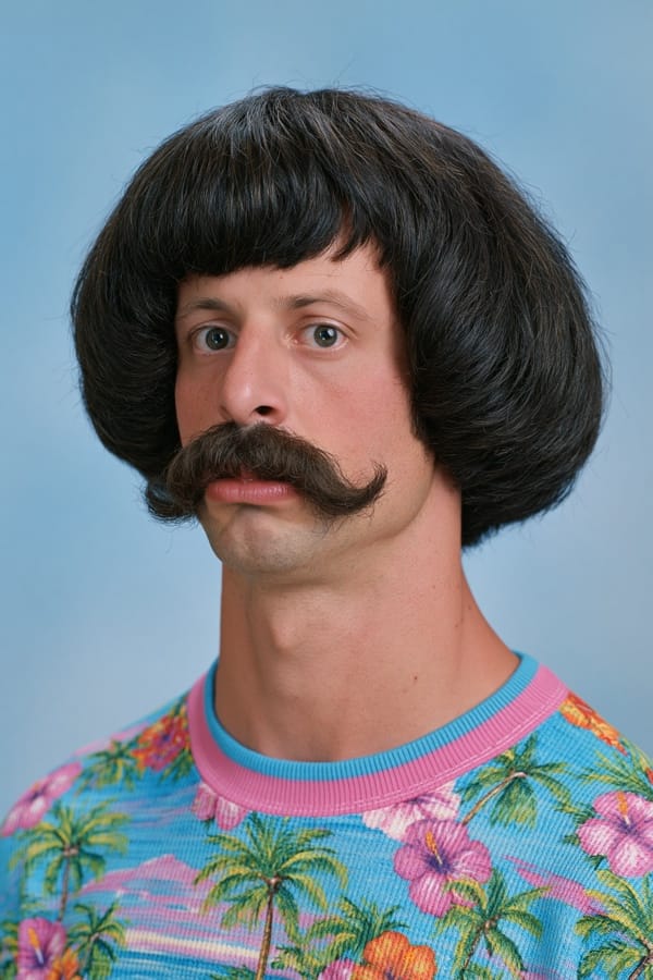 Studio yearbook portrait of a man with a heavy bowl haircut and a large handlebar moustache, wearing a bright tropical print sweatshirt against a pale blue backdrop