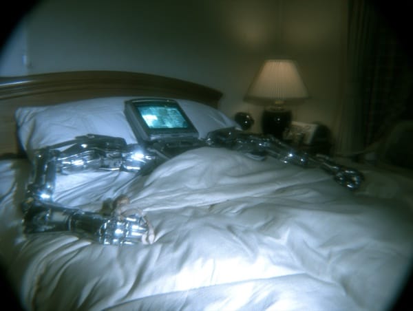 A handheld computer with a lit screen tucked under a duvet on a bed like a sleeping person, chrome robotic arms extending from its sides, one metal hand cradling a tiny human figure, dim bedside lamp burning