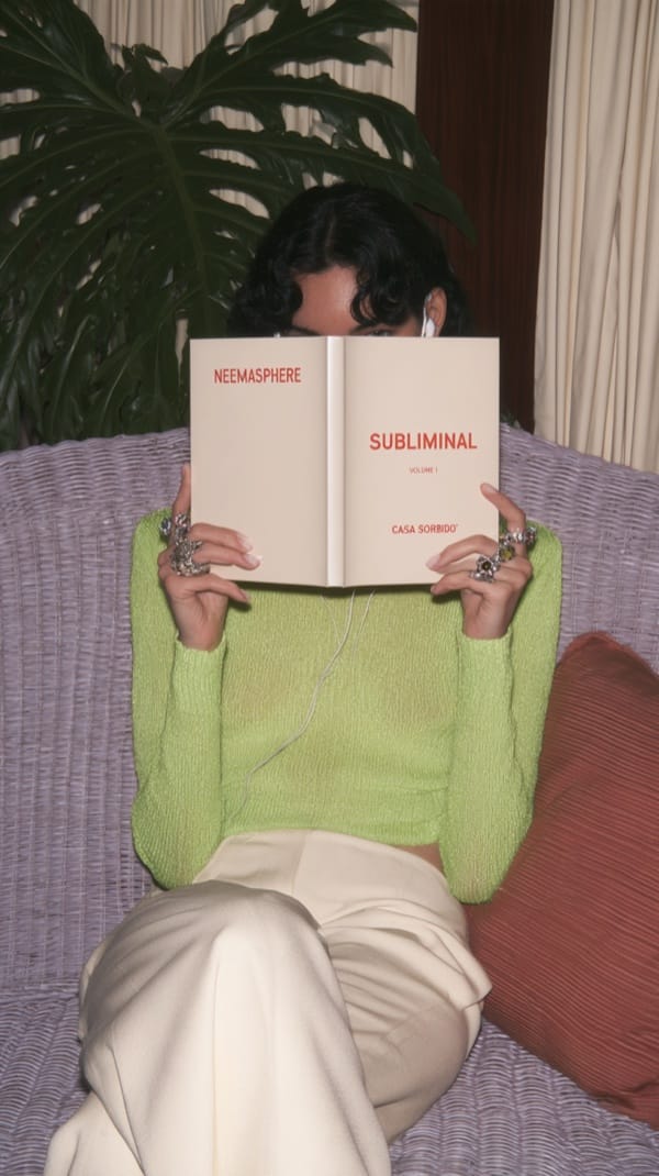 A person in a lime green knit top and cream trousers sits on a wicker bench, holding a cream book with red lettering in front of their face, beside a large leafy plant.