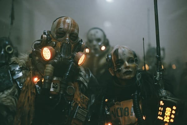 Explorers in rusted gas masks and scavenged gear crowd a fog-filled industrial hall, amber respirator filters glowing, a woman with a headset and pale cropped hair in front