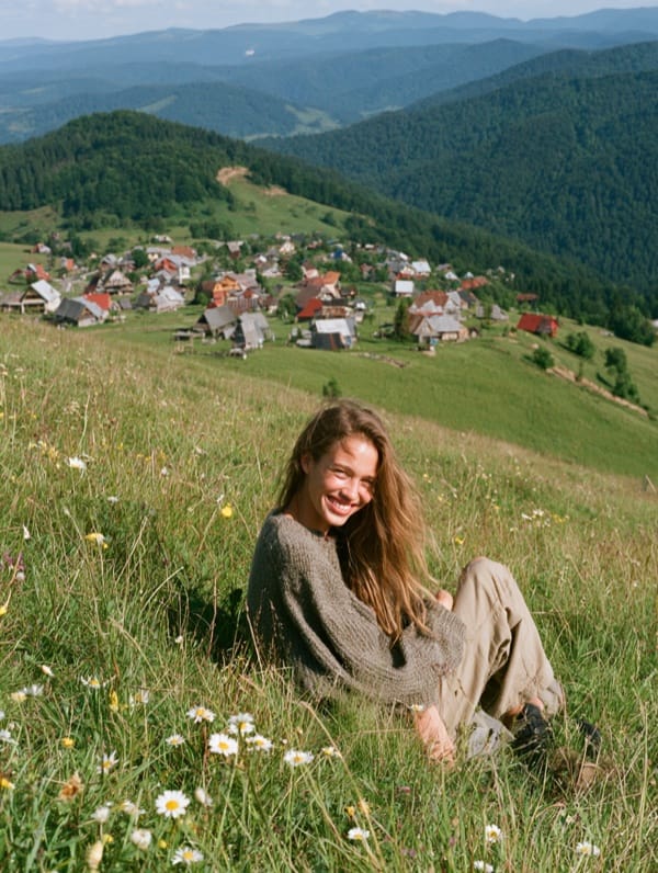 Photo-style image of a smiling young woman with long hair in a gray knit sweater, sitting in a grassy meadow with daisies on a hillside above a village of small houses, forested mountains behind